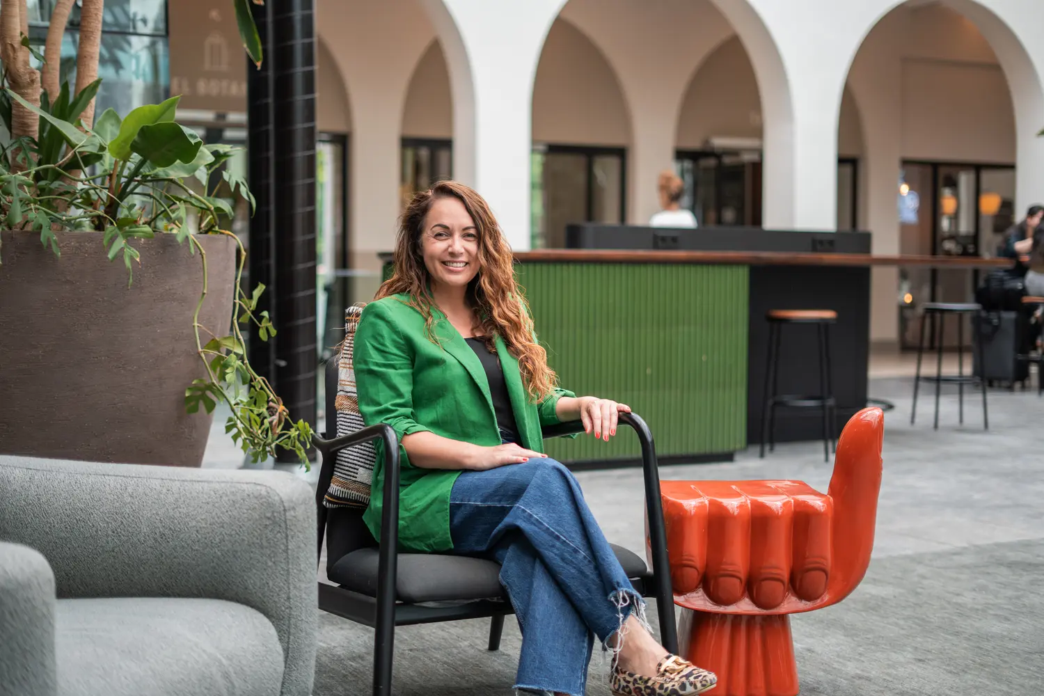 Vrouw in groene blazer zittend op een stoel in een modern kantoorinterieur met boogvormige doorgangen en groene accenten. Context: Coaching2Do, Meral Akar, Outplacement, Spoor 2 Begeleiding.