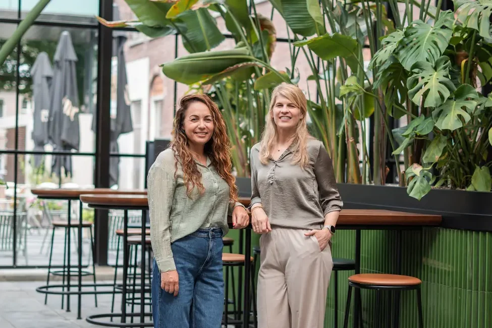 Twee vrouwen in een moderne kantoorruimte met veel groene planten, representerend Coaching2Do en Meral Akar, gespecialiseerd in outplacement en spoor 2 begeleiding.