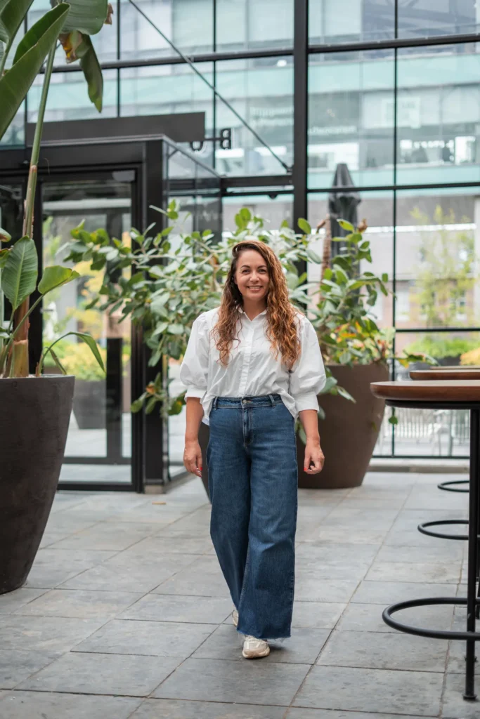 Woman walking in a modern indoor space with plants, wearing a white blouse and jeans, representing professional coaching and career guidance in a natural setting. Coaching2Do, Meral Akar, Outplacement, Spoor 2 Begeleiding.