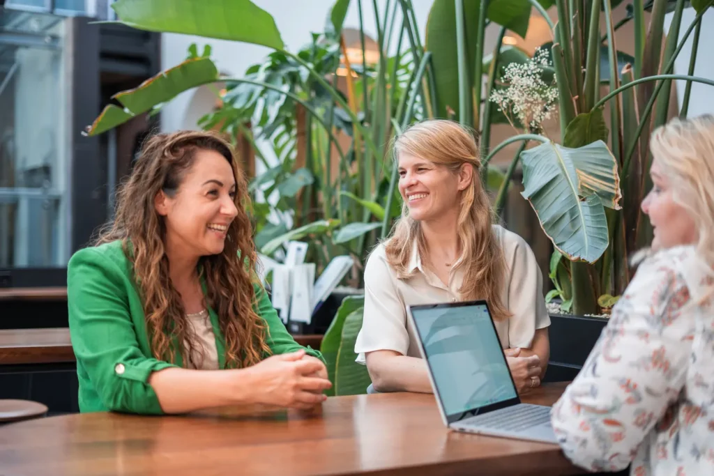 Drie vrouwen in gesprek aan tafel met laptop te midden van groene planten, voor Coaching2Do samenwerking en persoonlijke ontwikkeling.