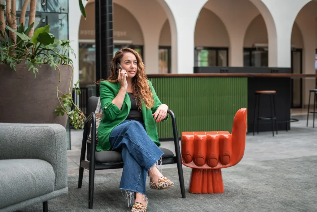 Vrouw in groene blazer voert telefonisch gesprek in moderne kantooromgeving naast een opvallende rode handstoel. Context: Coaching2Do, Meral Akar, Outplacement, Spoor 2 Begeleiding.