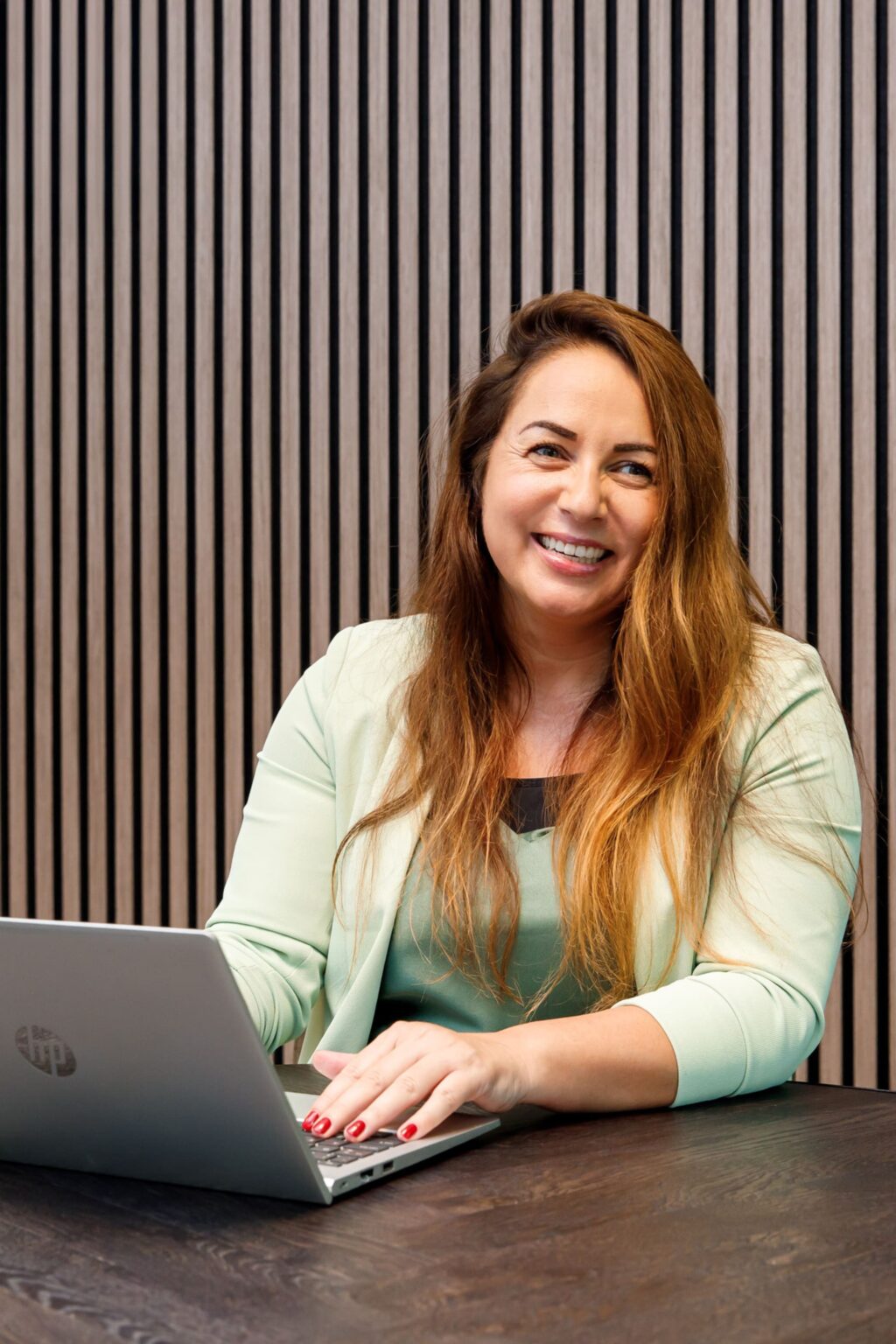 Glimlachende vrouw in een lichtgroene blazer werkt op een laptop met een houten lattenwand als achtergrond Meral Akar Persoonlijke coaching