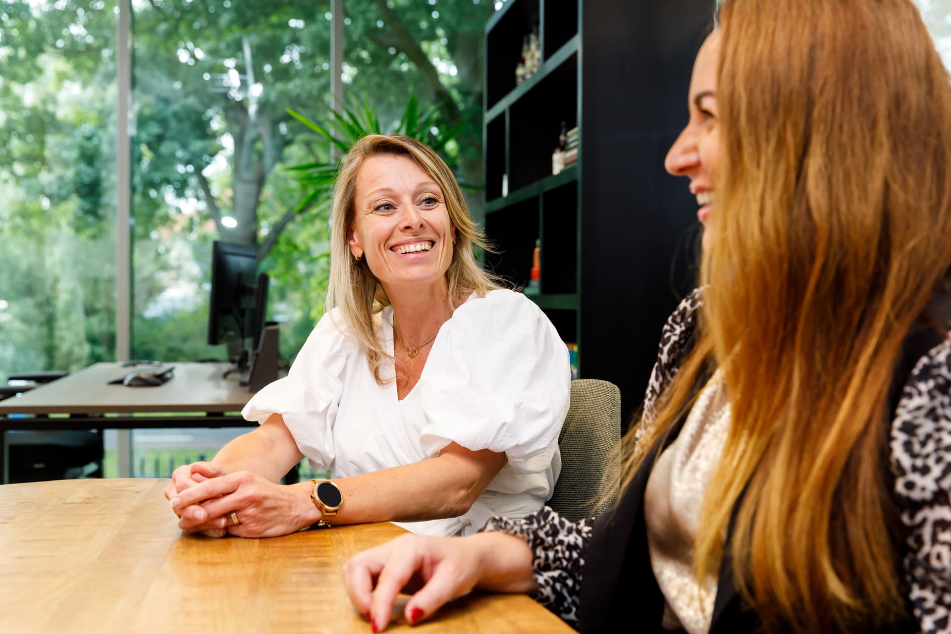 Lachende collega's in gesprek aan een houten tafel in een kantoor met groene omgeving over coaching2do