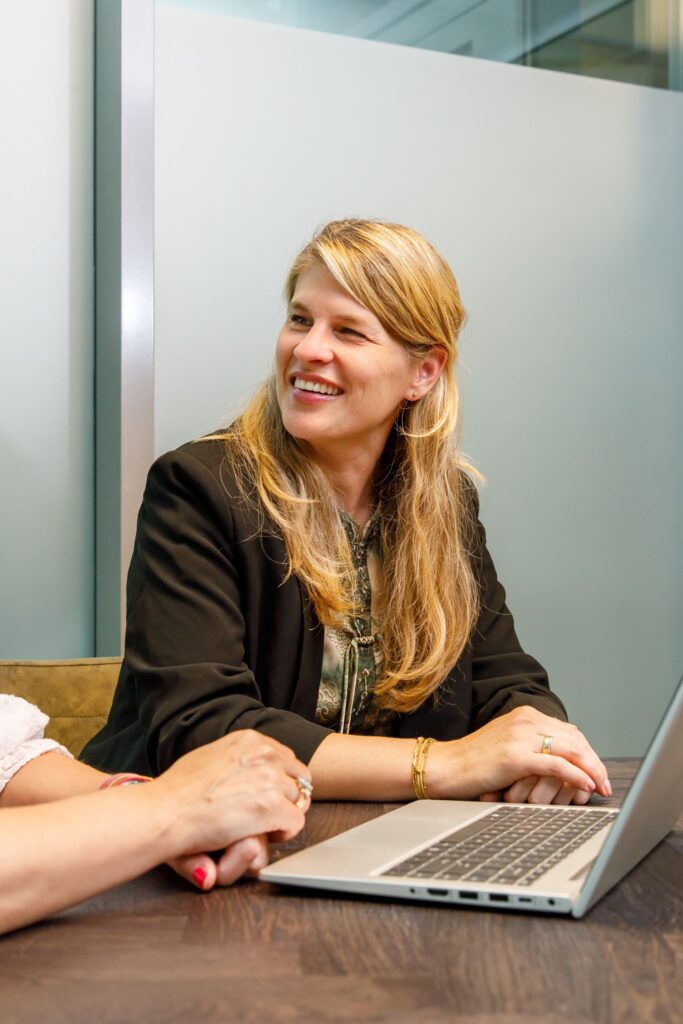 Vrouw in zwarte blazer lacht tijdens een zakelijke bijeenkomst met een laptop op een houten tafel