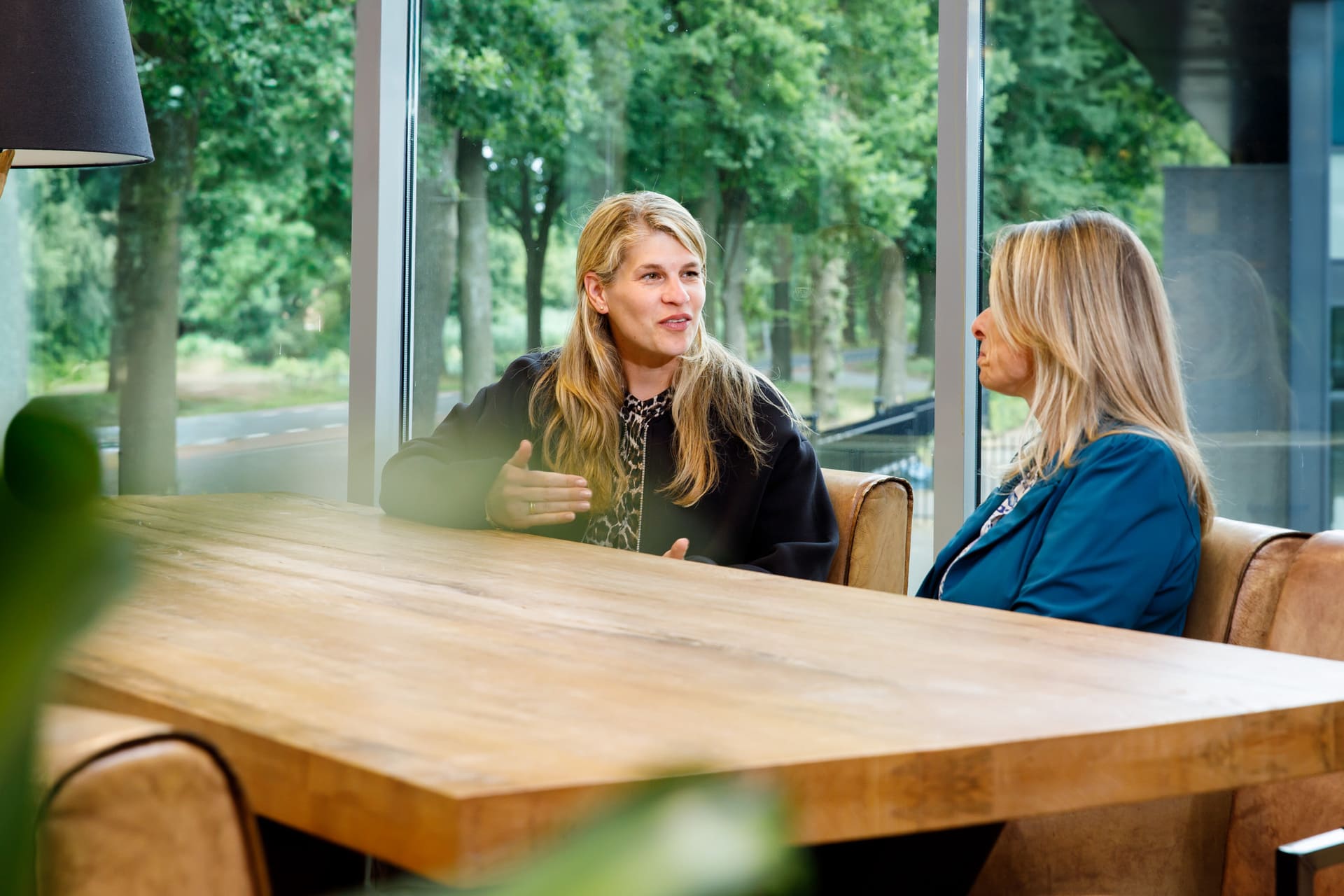 Twee vrouwen in gesprek aan een houten tafel met grote ramen, uitkijkend op een groene omgeving in gesprek over outplacement bij coaching2do